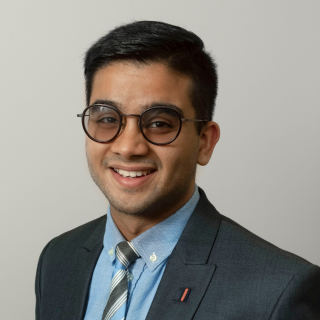 Abhinav is smiling at the camera wearing a blue suit and tie and black rimmed glasses.