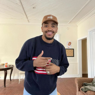 Amir is standing in an off white room with hardwood floors, smiling at the camera. He is wearing a sweatshirt with the American flag on the front and a tan hat.