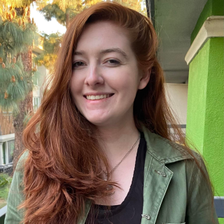 Emma is smiling at the camera wearing a green jacket and a black shirt. Her red hair is combed to one side.