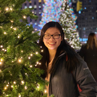 Jessica is standing next to a Christmas tree with lights smiling at the camera. She is wearing a dark grey jacket, glasses and her hair is parted just off to the side.
