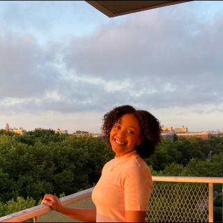 Yakira is standing in front of a tree canopy on a balcony with grey clouds in the sky. She is smiling at the camera with her body turned to one side and wearing a peach colored top. The sun is lighting her softly.
