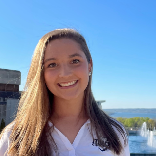 Megan is standing in front of the Ic Fountains with a blue sky behind her. She is smiling at the camera and wearing a white polo shirt.