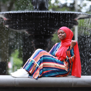 Khadija is sitting sideways in front of a large black fountain looking at the camera. She is wearing a bright red hijab and a multicolored stripped dress with white tennis shoes.