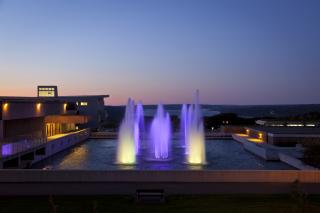 A view of the Ithaca College fountains and the lake beyond at sunset. The water in the fountains is colorfully lit and appears purple.