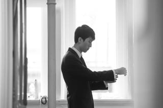 black and white photo of pianist Eric Lu standing in front of a bright window, adjusting the cuffs of a black jacket.