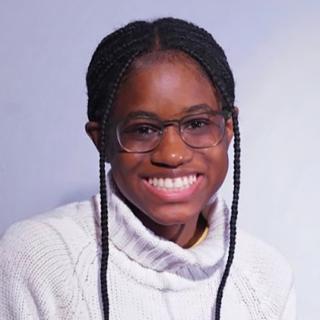 Alexus is starting directly into the camera for standard headshot on light grey/blue background. Alexus is wearing a white sweater and glasses, with two long braids framing each side of face. Alexus is smiling.