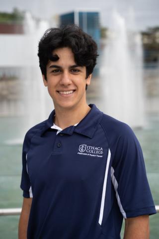 Alonso is posing for photo in front of Ithaca College Dillingham Fountains.  Alonso is wearing a IC physical therapy blue polo shirt. Is staring at the camera and smiling with black hair.