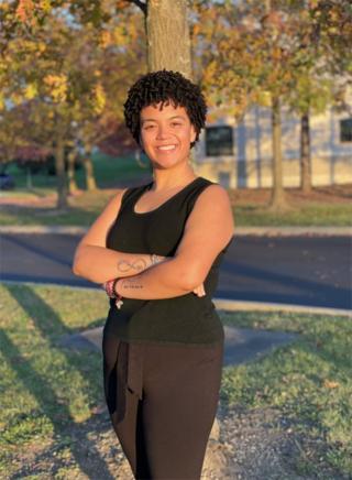 Briana is posing for photo with trees in background during autumn. Is wearing brown pants and a black sleeveless top. Has short hair. Is looking at camera and smiling.