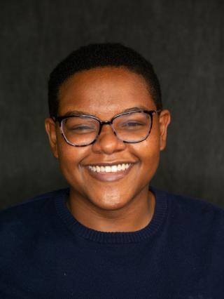 Brianna is posing for a camera headshot on black background. Wearing a black shirt and block rimmed glasses.  Is smiling.