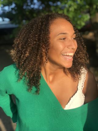 Cayla is staring off to her right and is smiling. She is wearing a green sweater with a white tank top exposed over her left shoulder.