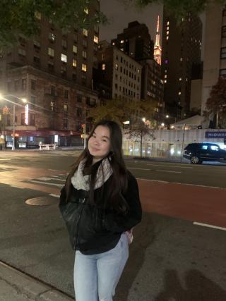 Frances is posing for photo in urban setting at night. Is wearing light jeans and black coat with white scarf. Has long dark hair extending past shoulders.