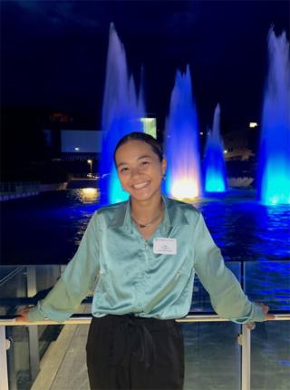 Irena is posing in front of the Dillingham Fountains lit blue at night. Is wearing seafoam green shirt and black pants. Is smiling at camera.
