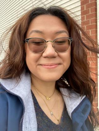 Isabella is posing in front of building with off-white vinyl siding. Is wearing grey shrit with blue jacket. Is wearing glasses and multiple necklaces. Is smiling and has shoulder length brown hair.