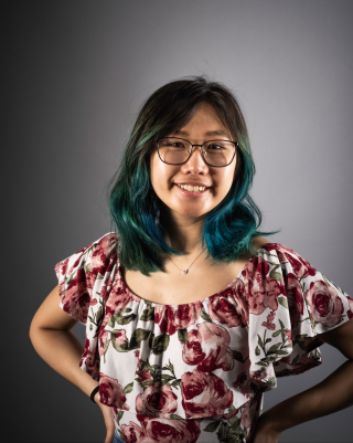 Jaquelyn is posing for standard headshot on dark grey background. Wearing red and white floral patterned top and glasses. Is smiling.