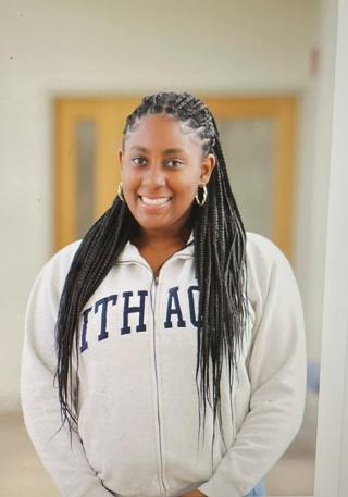 Jolivia is posing for picture in classroom with white walls and light wooden door in background. Is wearing a white Ithaca zip up sweater with blue lettering. Has dark braided hair extending well past shoulders. Jolivia is staring at camera and smiling.