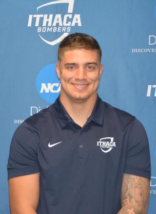 Joseph is posing for photo in front of blue background with Ithaca and NCAA logos. Is wearing a blue polo shirt. Has a tattoo on left arm. Has short brown hair. Is looking directly at the camera and smiling.