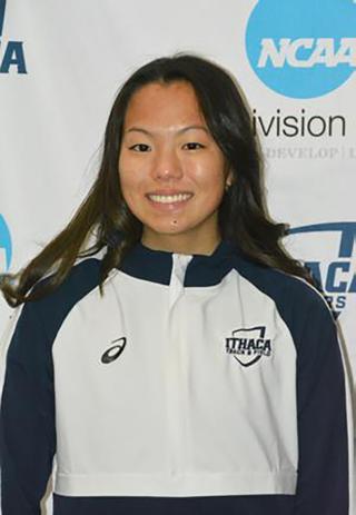Julia is posing for headshot on white background with NCAA and Ithaca College logos. Is wearing a white and blue Ithaca College track and field jacket. Is smiling and looking at camera. Has dark hair extending past shoulders.