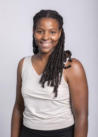 Kalena is posing for picture on light grey/white background. Is wearing a white top with no sleaves. Kalena is looking at camera and smiling. Has dark brown braided hair extending past shoulders.