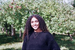 Martha is posing in orchard with apple trees in background. Is wearing a black sweater and gold necklace. Has shoulder length dark curly hair. Is smiling directly at camera on sunny day.