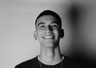 Mateo is posing for headshot on light background. Is black and white photo. Is wearing a black top and necklace. Is looking at camera and smiling with high fade hair cut.