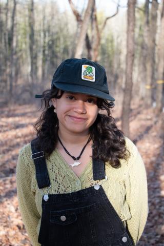 Melanie is posing for camera with barren forest in background. Wearing a green sweater and black overalls and a black hat with logo. Has shoulder length dark hair.