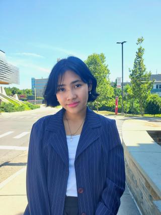 Nano is posing for photo with road and buildings and tree in background. Is wearing a white top with blue striped blazer. Is wearing a necklace. Has short dark hair.
