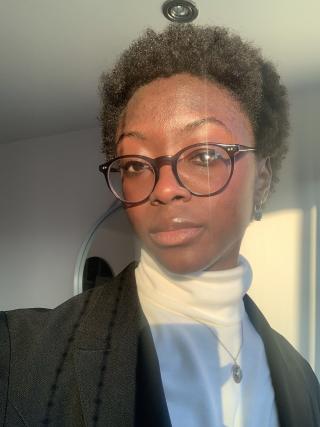 Nijha is posing for camera in room with white walls. Is wearing a white turtleneck sweater and black dress coat and glasses.