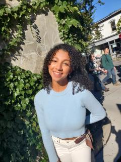 Stephanie is posing for photo in front of brick wall covered in green ivy. Serindipity building is in the background as well as two bypassers. Stephanie is wearing light blue sweater and white pants. Has dark curly hair extending to shoulders. Is looking at camera and smiling.