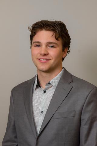 Tyler is posing for photo in front of off-white background. Is wearing a light grey shirt with dark grey blazer. Has short hair. Is looking at the camera and smiling.