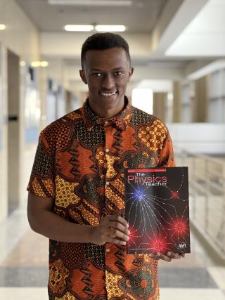 Ted Mburu stands in a hallway in the physics building holding a copy of The Physics Teacher.