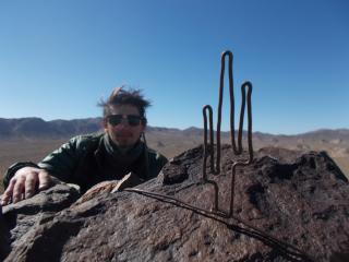 Porter is posed for a photo in a desert setting. Porter has hand on a rock and has a barren background with blue sky. Porter is wearing sunglasses. There is a metal object in the shape of a cactus.