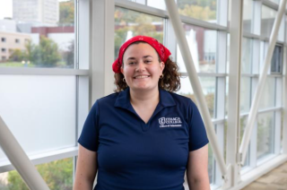 Skyler is posing for photo in Ithaca College walkway. Skyler is wearing blue Ithaca polo shirt and a red bandana. Skyler is facing the camera and is smiling.