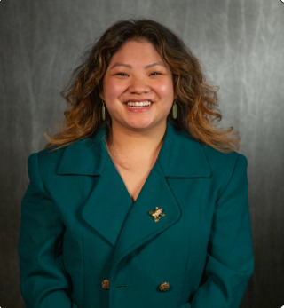 Kristin is posing for photo on grey patterned background. Is wearing green overcoat with gold pins. Has brown shoulder length hair and is smiling at camera.