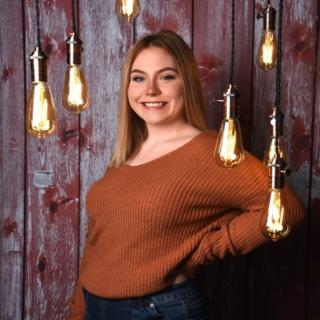 Katelynn is posing for photo in front of a wooden plank background with lighting fixtures hanging down. Katelynn is wearing a burnt orange top. Katelynn is looking at the camera and smiling.