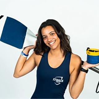 Nicole is wearing a rowing wet suit and is holding an oar behind head. Nicole is staring at the camera and is smiling.