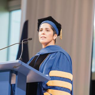 A woman in academic cap and gown stands at a podium with microphones and looks out over the crowd. 
