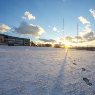 Winter sunset over footprints in a snow-covered field with a campus building in the far back. 