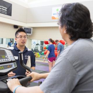 A student in a blue shirt wearing glasses is observing a woman in a gray shirt that is on a treadmill. In the background, there are two people doing exercises.