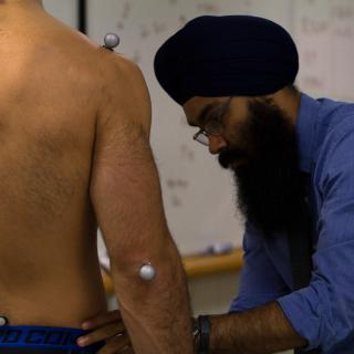 A man in a blue shirt is placing sensors on a man without a shirt on along various muscles so that he can measure his movements when he begins to run.