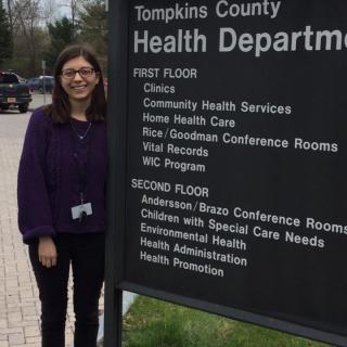 This is a picture of a student standing next to the Tompkins County Health Department sign which lists the different types of services offered such as clinics, immunizations, and vital records.