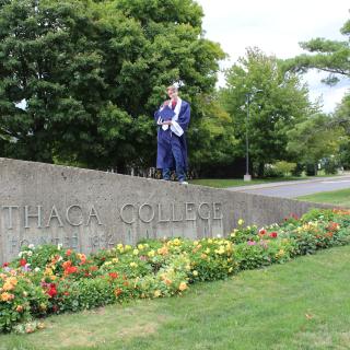 Person standing in graduation regalia on top of stone sign reading "Ithaca College"
