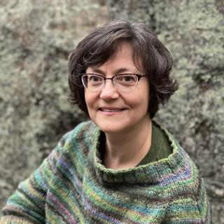 white woman with glasses and brown hair sitting in front of nature. different hues of green striped sweater.