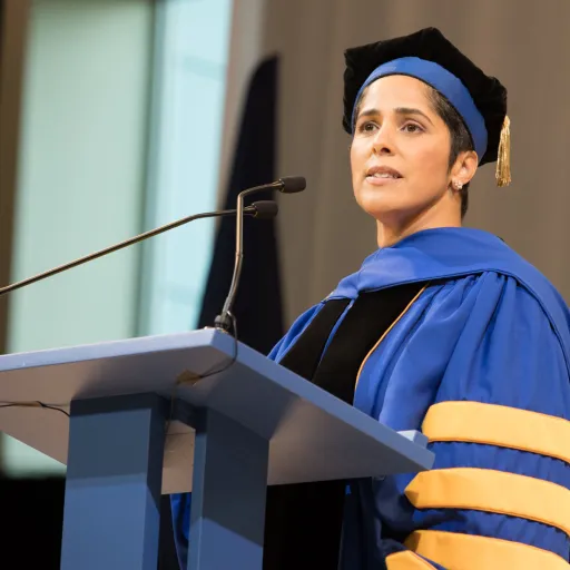 A woman in academic cap and gown stands at a podium with microphones and looks out over the crowd.