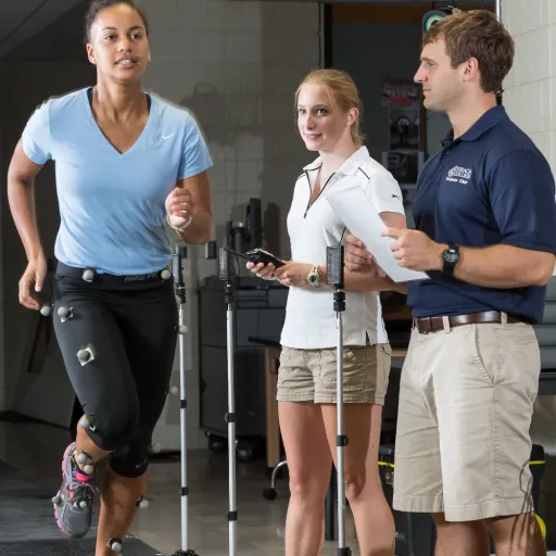 This is a picture of an athlete with a blue shirt and black pants  running in the biometrics lab with sensors attached to the lower part of the body. Two students observe and are taking notes about what they are seeing while the athlete is running.