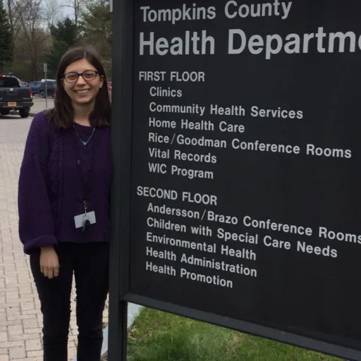 This is a picture of a student standing next to the Tompkins County Health Department sign which lists the different types of services offered such as clinics, immunizations, and vital records.
