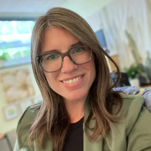 white woman with brown hair glasses is wearing a green blazer and black undershirt. background is a blury window, plant, and flower photo