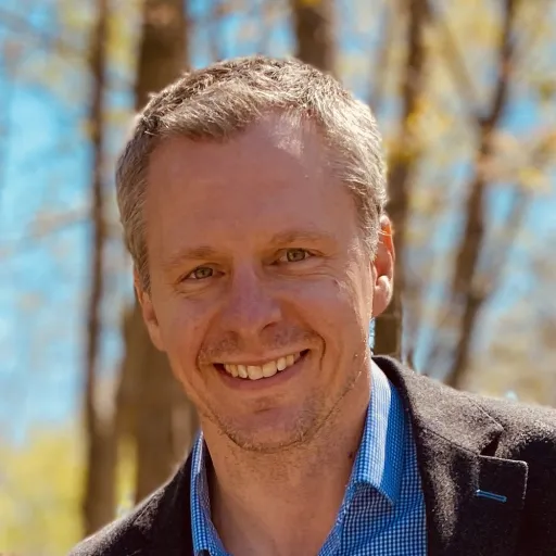 white male with blue collar and brown jacket smiling with trees in the background