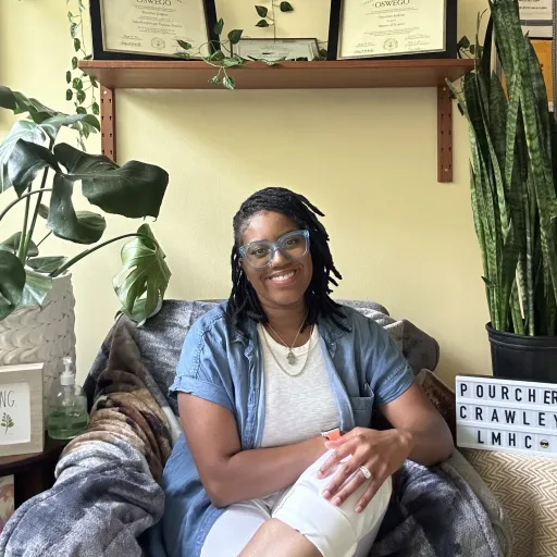 A black woman wearing a blue shirt with a white undershirt sits cross legged. she is surrounded by plants.