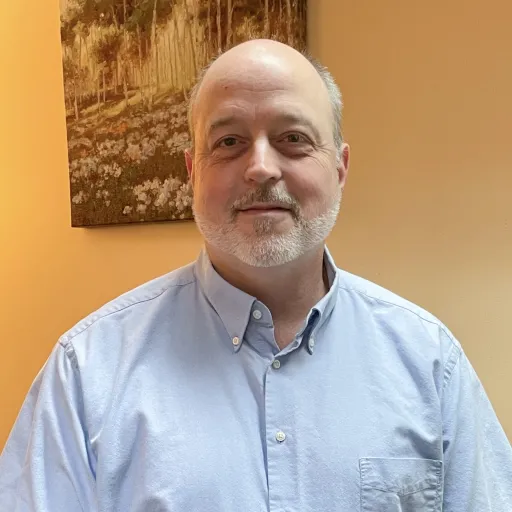 Brian is wearing a light blue color shirt, is grinning and standing in front of a painting
