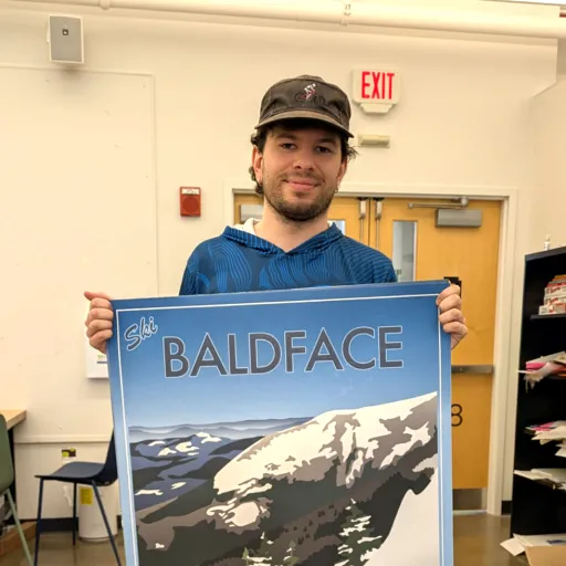 Adams Staples holds a large travel-style poster he designed, showing a skier in a red jacket on a snowy slope with the text "Ski Baldface, Chatham, New Hampshire."